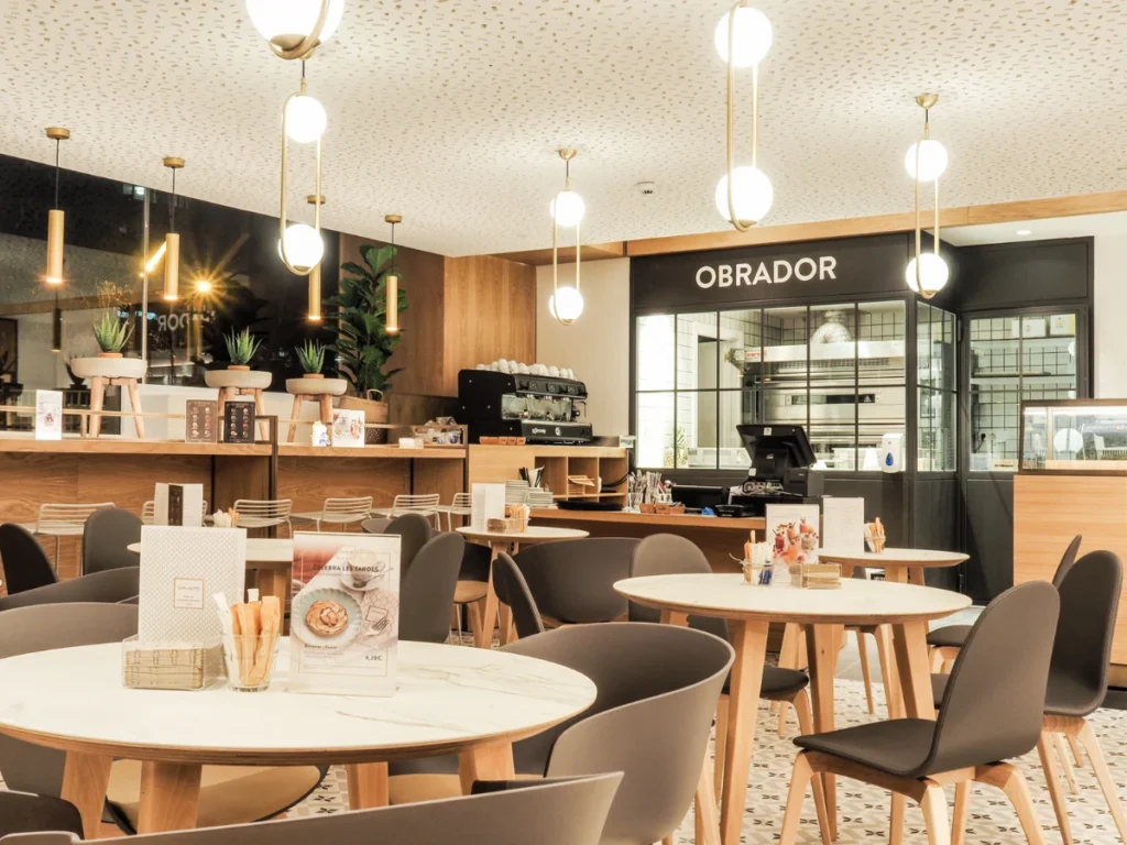 Sala de la cafetería Crustó Diagonal en Barcelona, con mesas, lámparas y el obrador al fondo