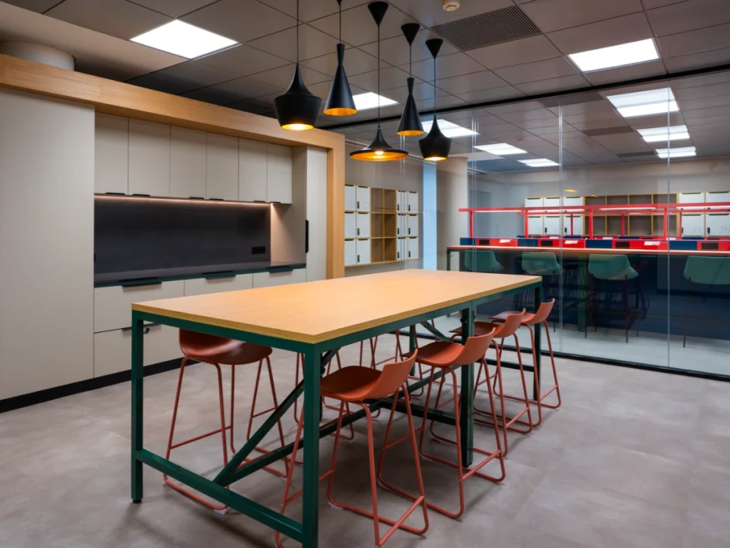 Zona de cocina con mesa de madera y taburetes, armarios y lámparas colgantes en las oficinas de Cheil